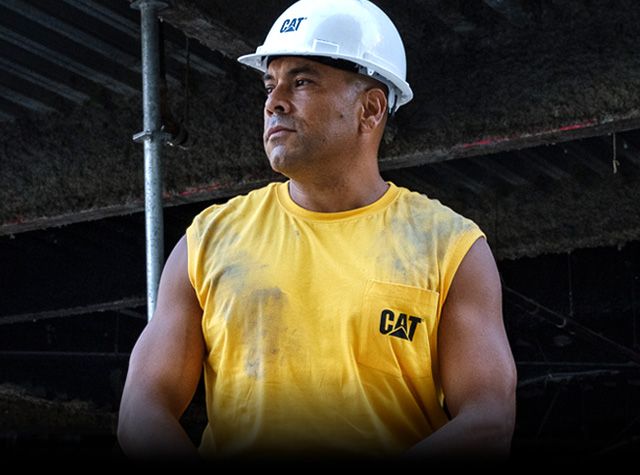 Man under scaffolding wearing a hard hat and a yellow CAT t-shirt.