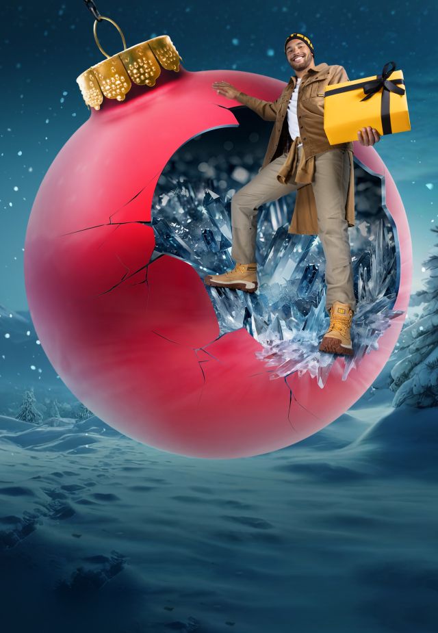 A person holiding a wrapped gift is standing on a large tree decoration suspended from heavy equipment in a wintry landscape.